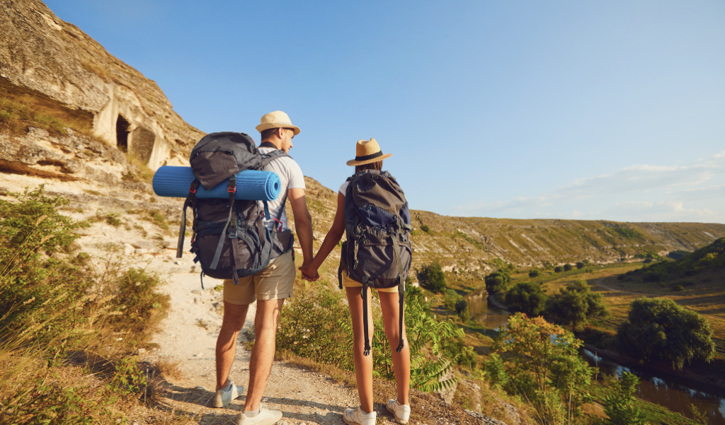 Two hikers carrying backpacks holding hands while walking on a trail.